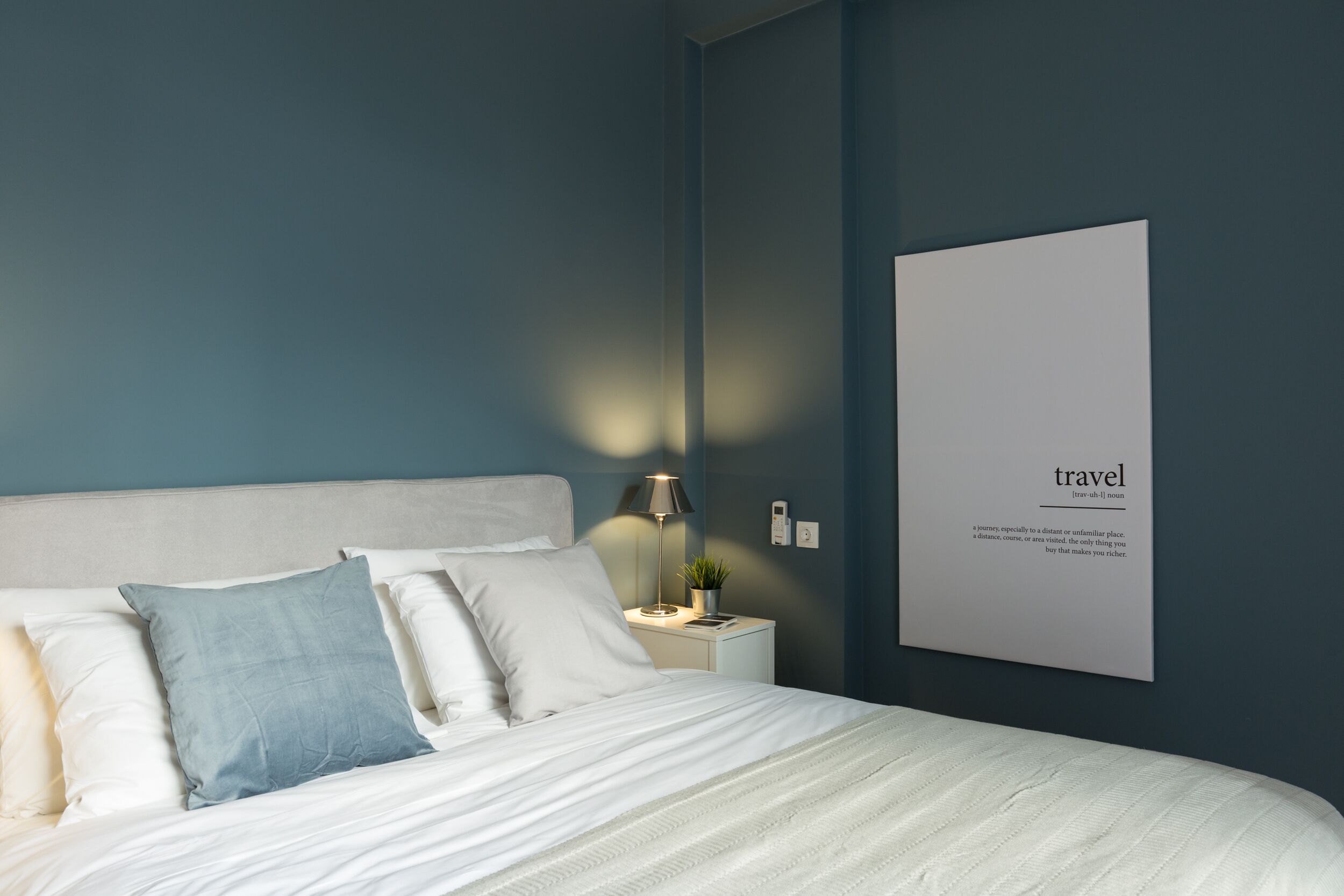 Una cama bien hecha con almohadas blancas y azules, una lámpara de noche y un texto enmarcado en la pared.