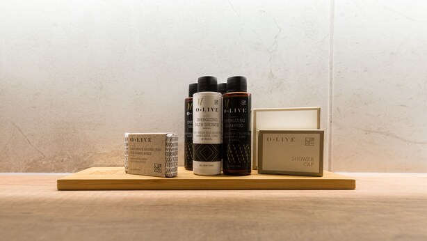 Une étagère en bois sur laquelle sont disposés divers produits de la marque O.L.I.V.E, notamment un bonnet de douche, des bouteilles de shampoing et de gel douche, ainsi qu’une boîte de savon naturel.