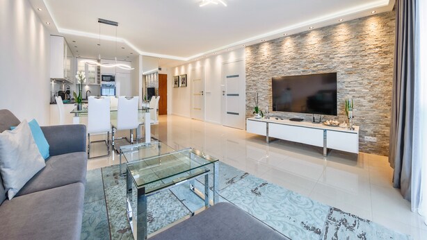 A modern living room with a glass coffee table, a sofa, a TV mounted on a stone wall, and a dining area in the background. - Apartamenty Sun & Snow Zwyciestwa (Miedzyzdroje)