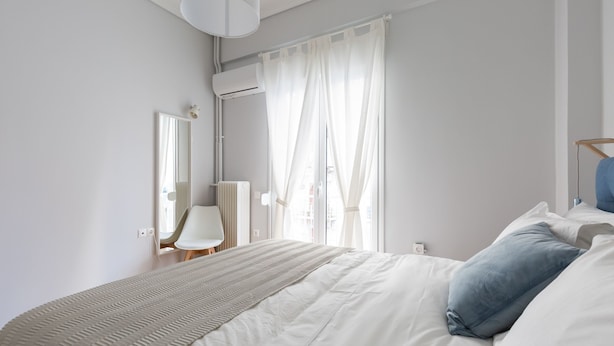 Um quarto moderno com uma cama grande, uma mesinha de cabeceira, um espelho e uma janela com cortinas leves.