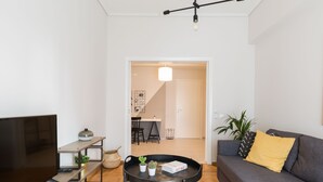 Uma sala de estar moderna com televisão de tela plana, uma mesa de centro com plantas, um sofá cinza com almofadas amarelas e um espaço de jantar ao fundo.