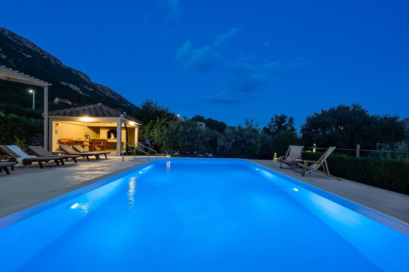 A swimming pool with lounge chairs and a covered seating area, surrounded by trees and mountains.