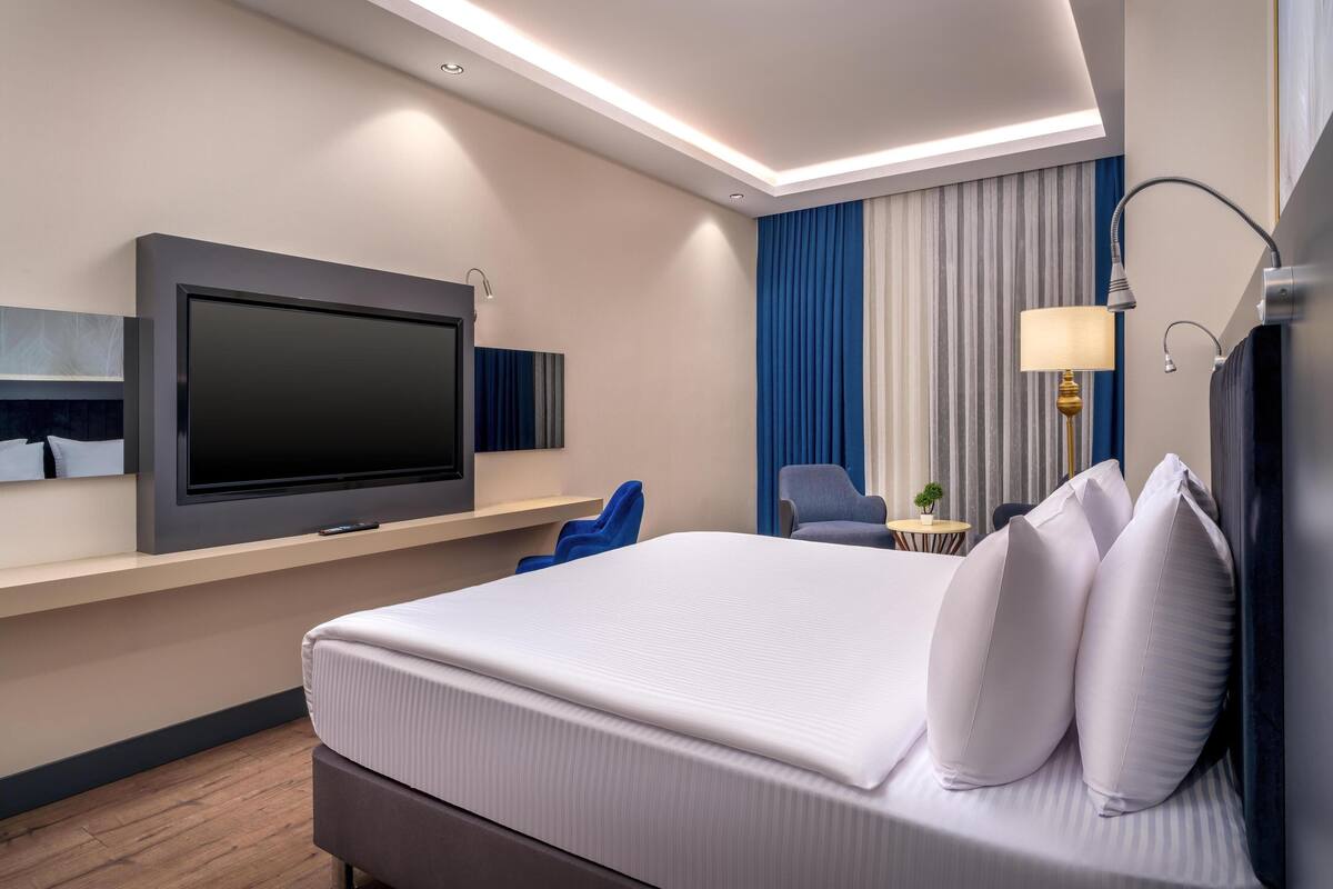 Une chambre d’hôtel moderne dotée d’un grand lit, d’un téléviseur à écran plat fixé au mur, d’un bureau avec une chaise et d’une petite table avec une lampe.