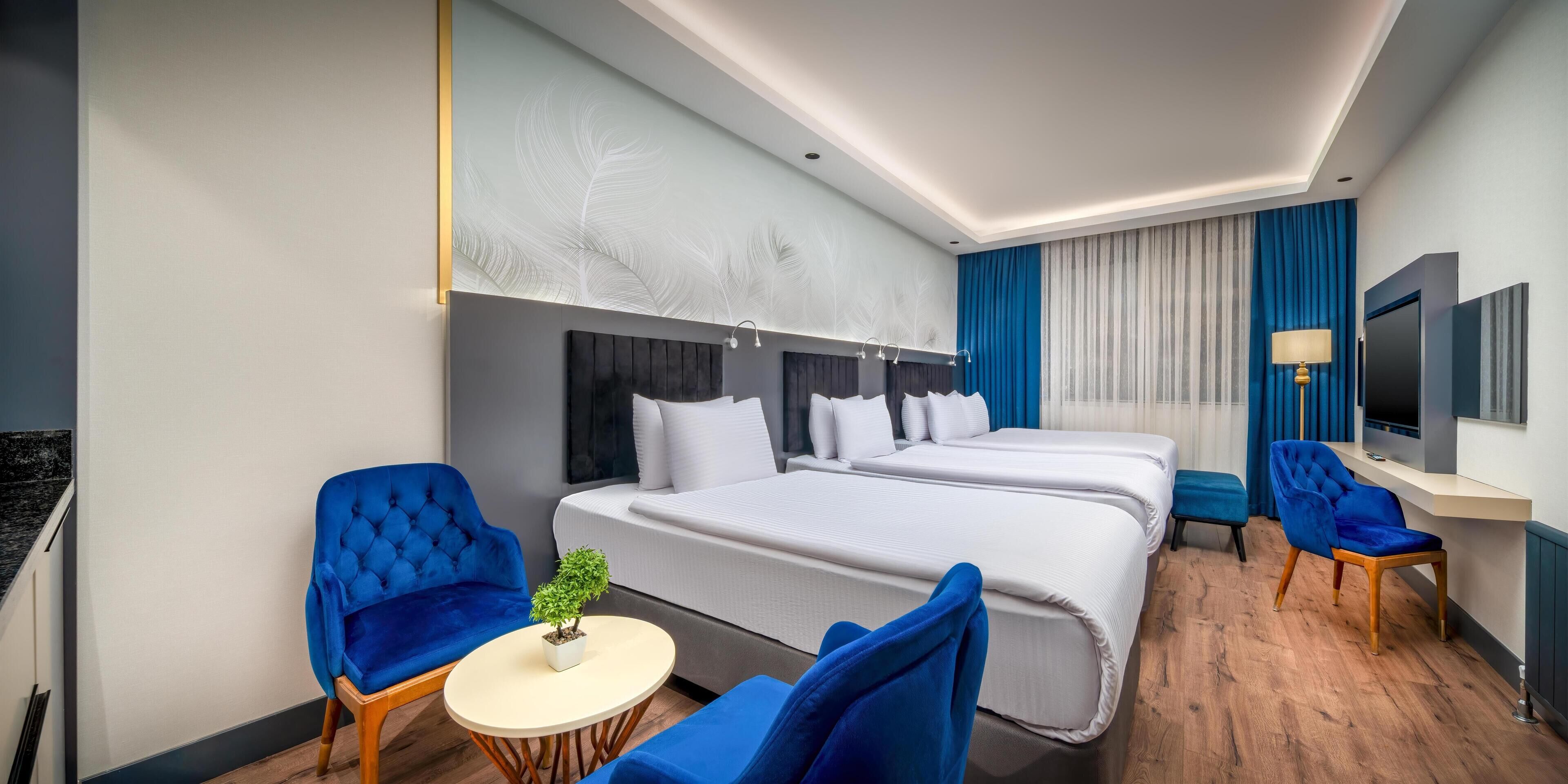 A hotel room with two beds, a small round table with a plant, two blue armchairs, a TV, and a desk with a lamp.