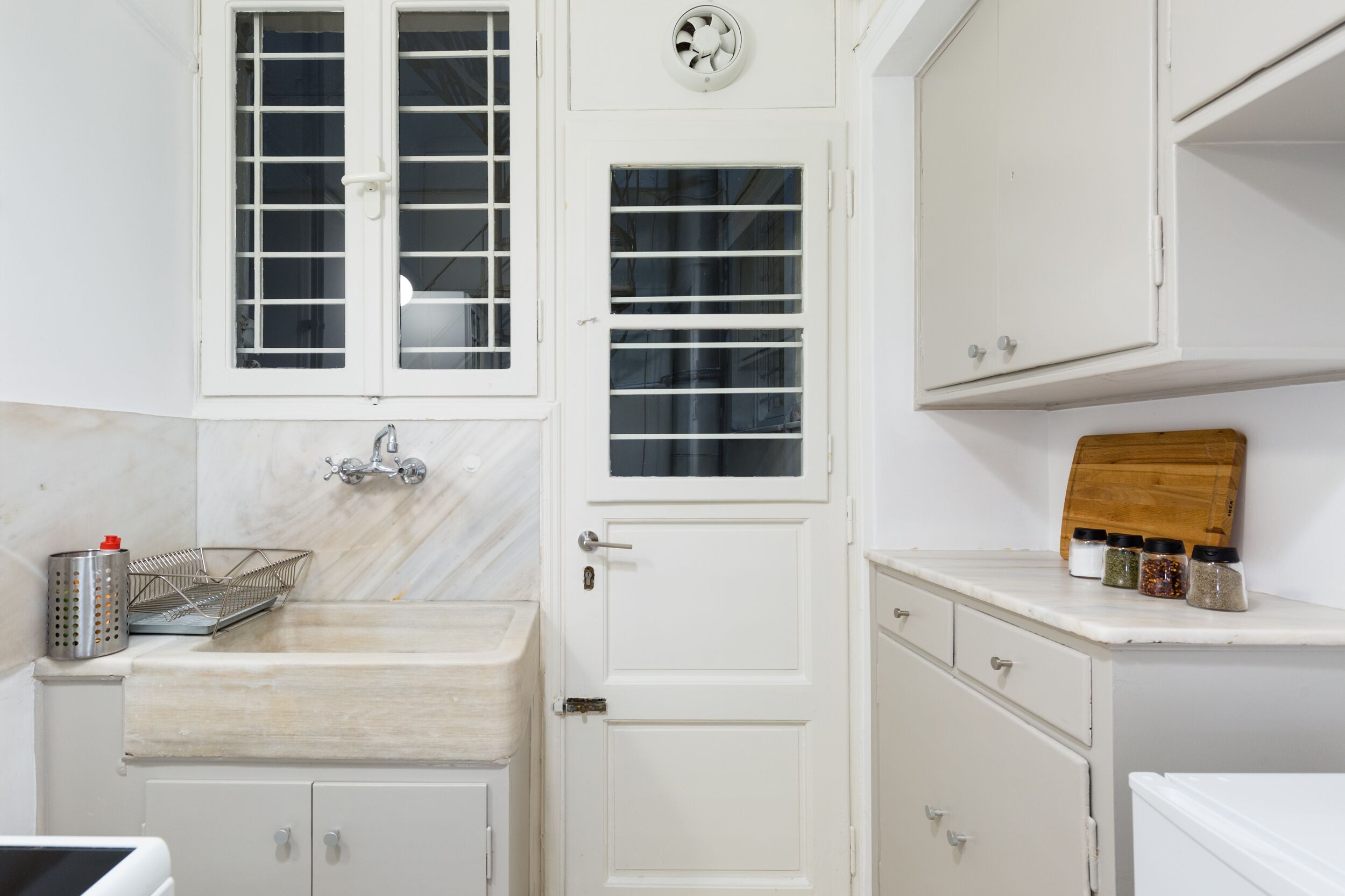 A kitchen with white cabinetry, a sink, a window with blinds, and a wooden cutting board with spices.