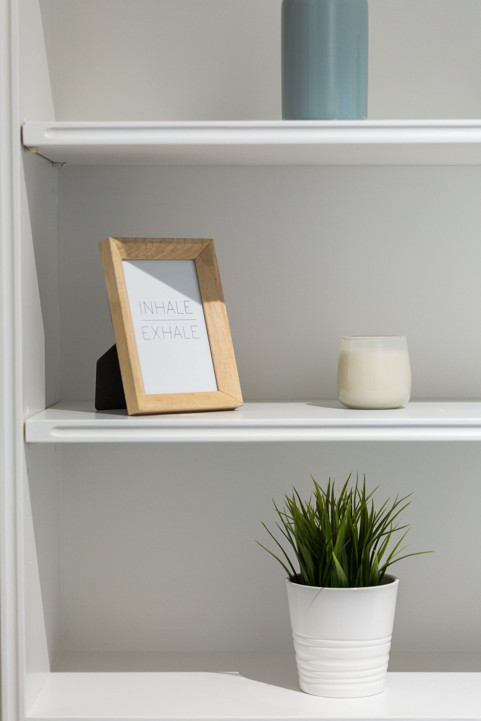 Uma prateleira branca com uma placa emoldurada, uma vela e um vaso de planta.