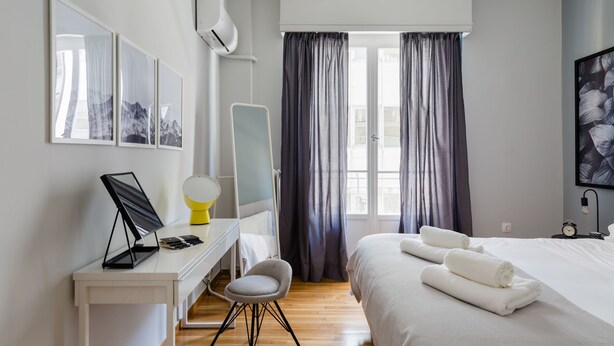 A modern hotel room with a bed, a desk with a chair, a bedside table, and a large window with curtains. - UPSTREET Acropolis Heart Apartments (Athens)