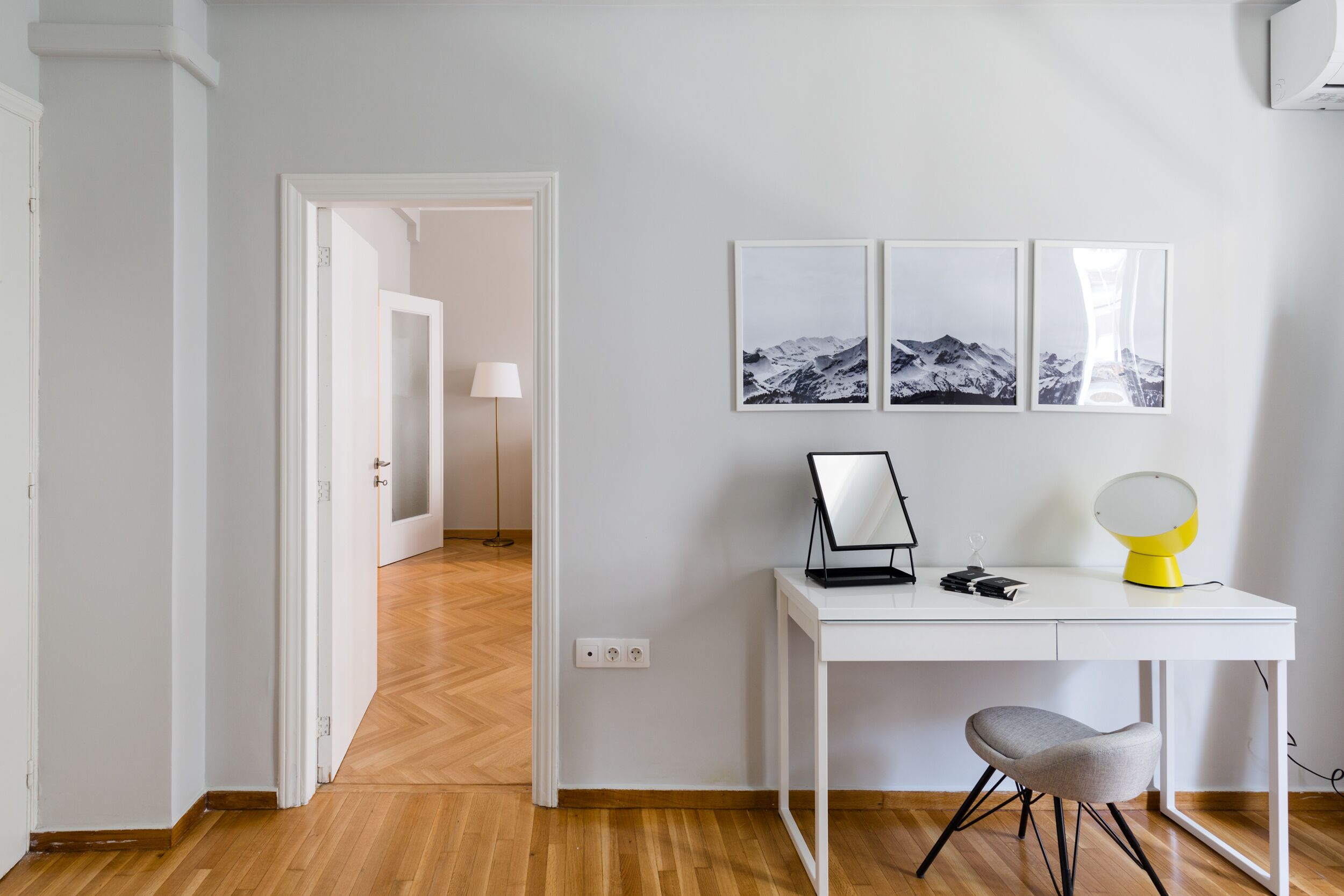 A modern room with a white desk, a chair, and a lamp. There are three framed pictures on the wall.