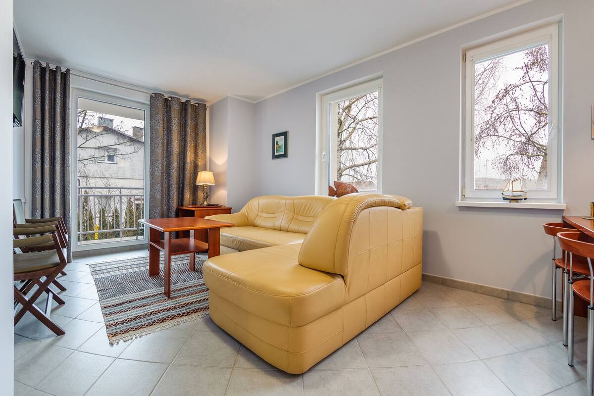 A living room with a sofa, a dining table with chairs, a small side table, a lamp, and a view of the outside through a window.