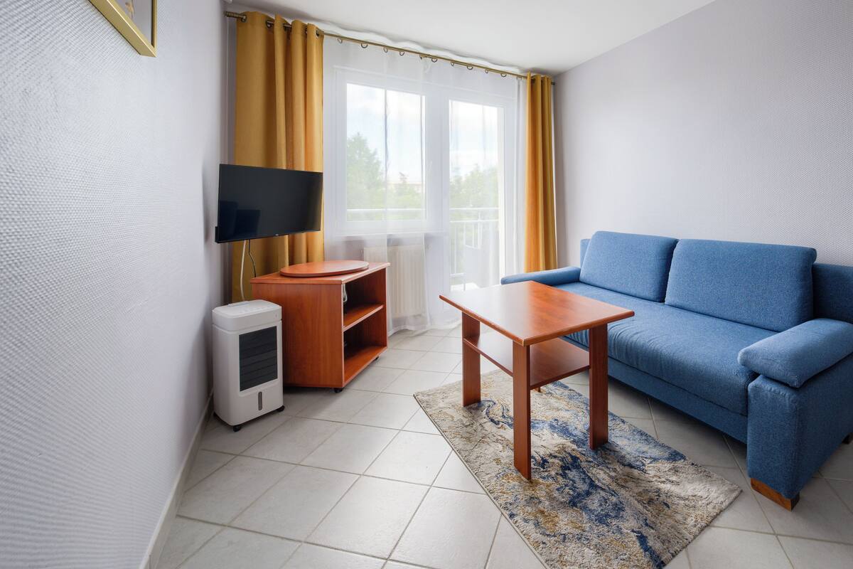 A modern living room with a blue sofa, a wooden coffee table, a TV mounted on the wall, and a small wooden side table.