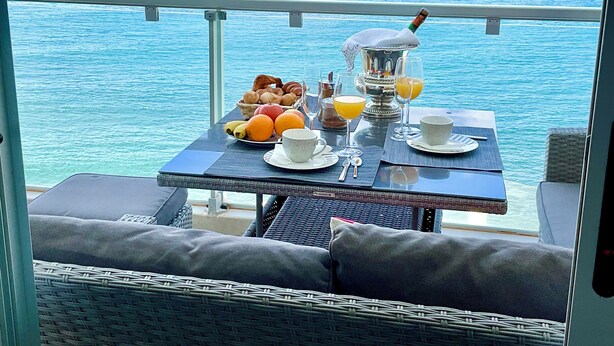 Een balkon met een tafel, ontbijtartikelen, een fles en uitzicht op zee.