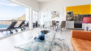 Een moderne woonkamer met een glazen salontafel, een bank en een eethoek met stoelen. Er is een keuken met een magnetron en oven, en een uitzicht op de oceaan door grote ramen.