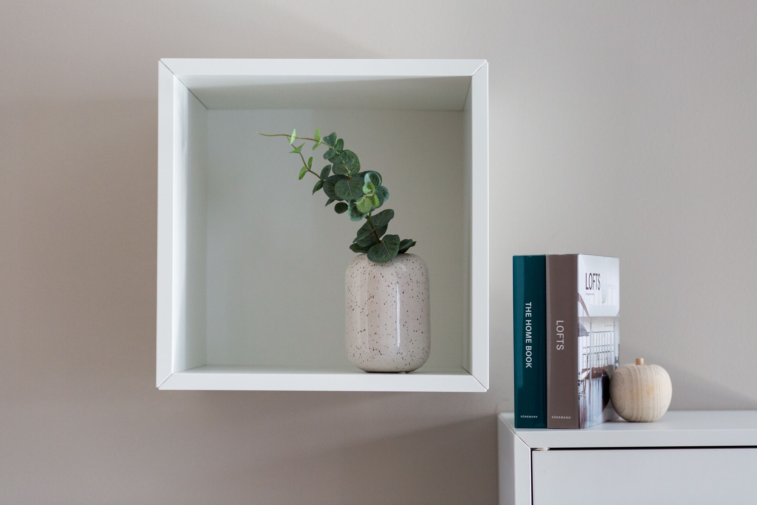 A white shelf with a ceramic vase containing a plant, a book titled 'The Home Book', and a small pumpkin.