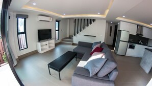 Una sala de estar moderna con un sofá gris, una mesa de centro negra y un televisor de pantalla plana. Se ve una escalera que lleva a un piso superior, y un área de cocina con armarios blancos y un refrigerador.
