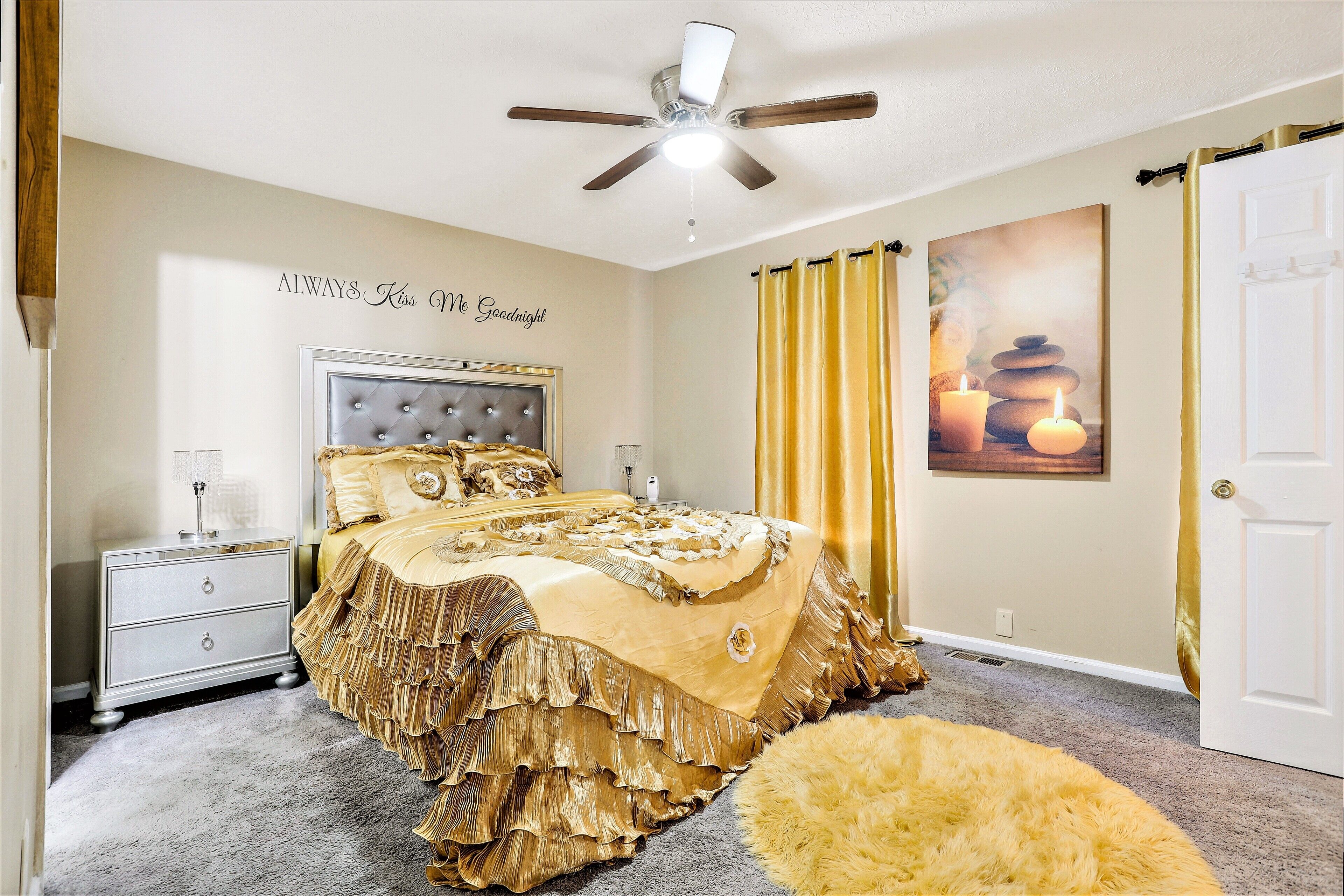 A bedroom with a tufted headboard, a bed with a ruffled gold bedspread, a nightstand with a lamp, and a ceiling fan.