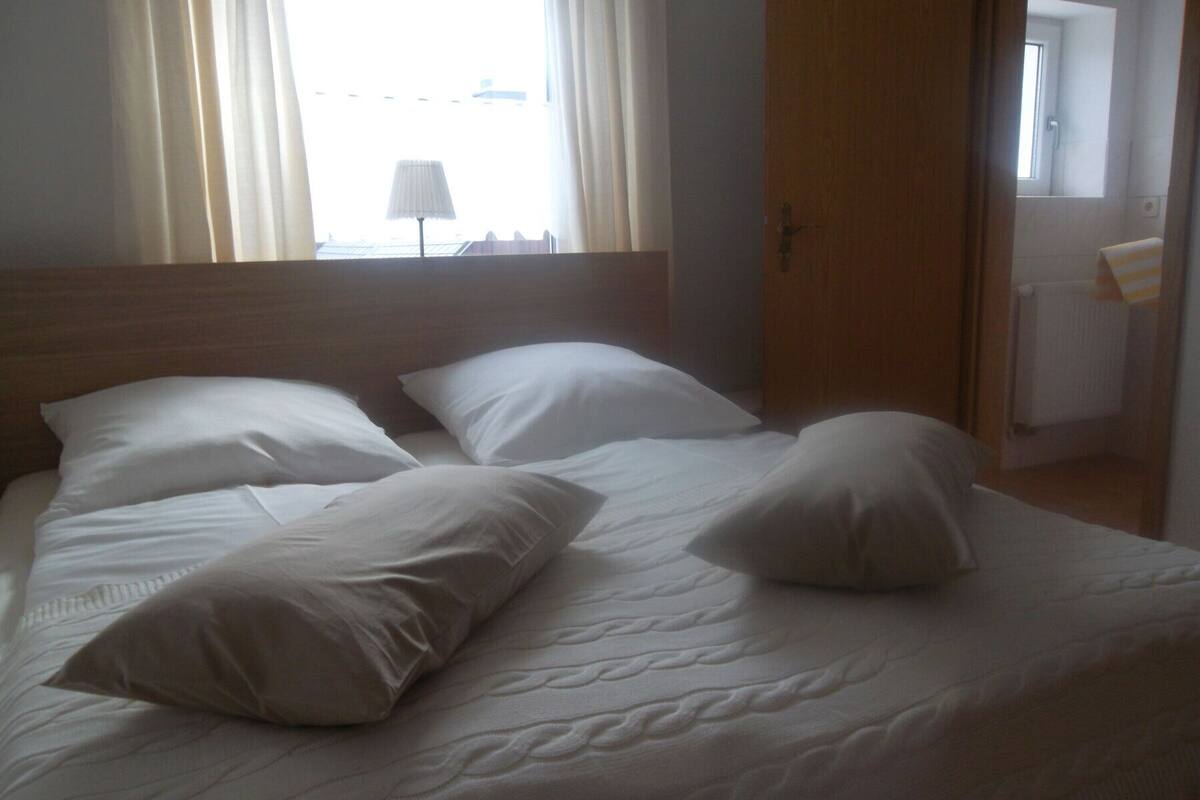 Una cama con ropa de cama y almohadas blancas, un cabecero de madera y una ventana con cortinas.