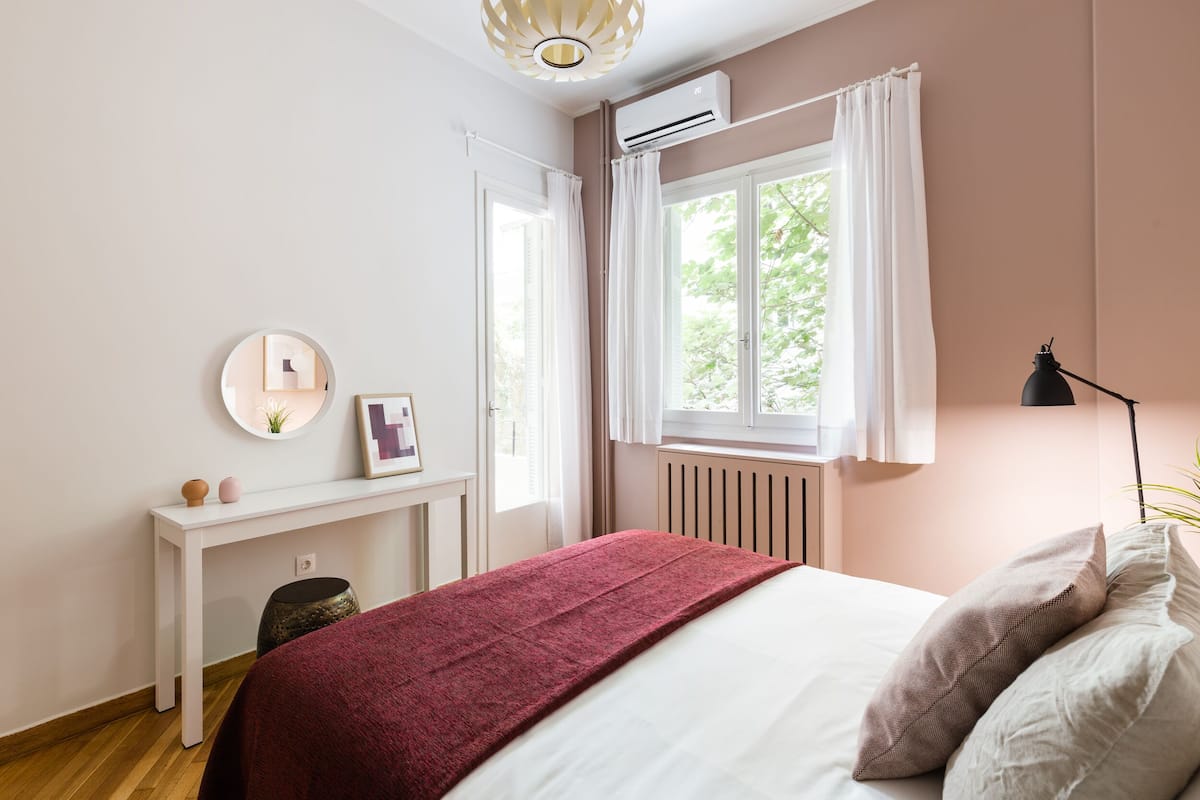 Un dormitorio con cama, mesita de noche, espejo, ventana con cortinas y aire acondicionado.
