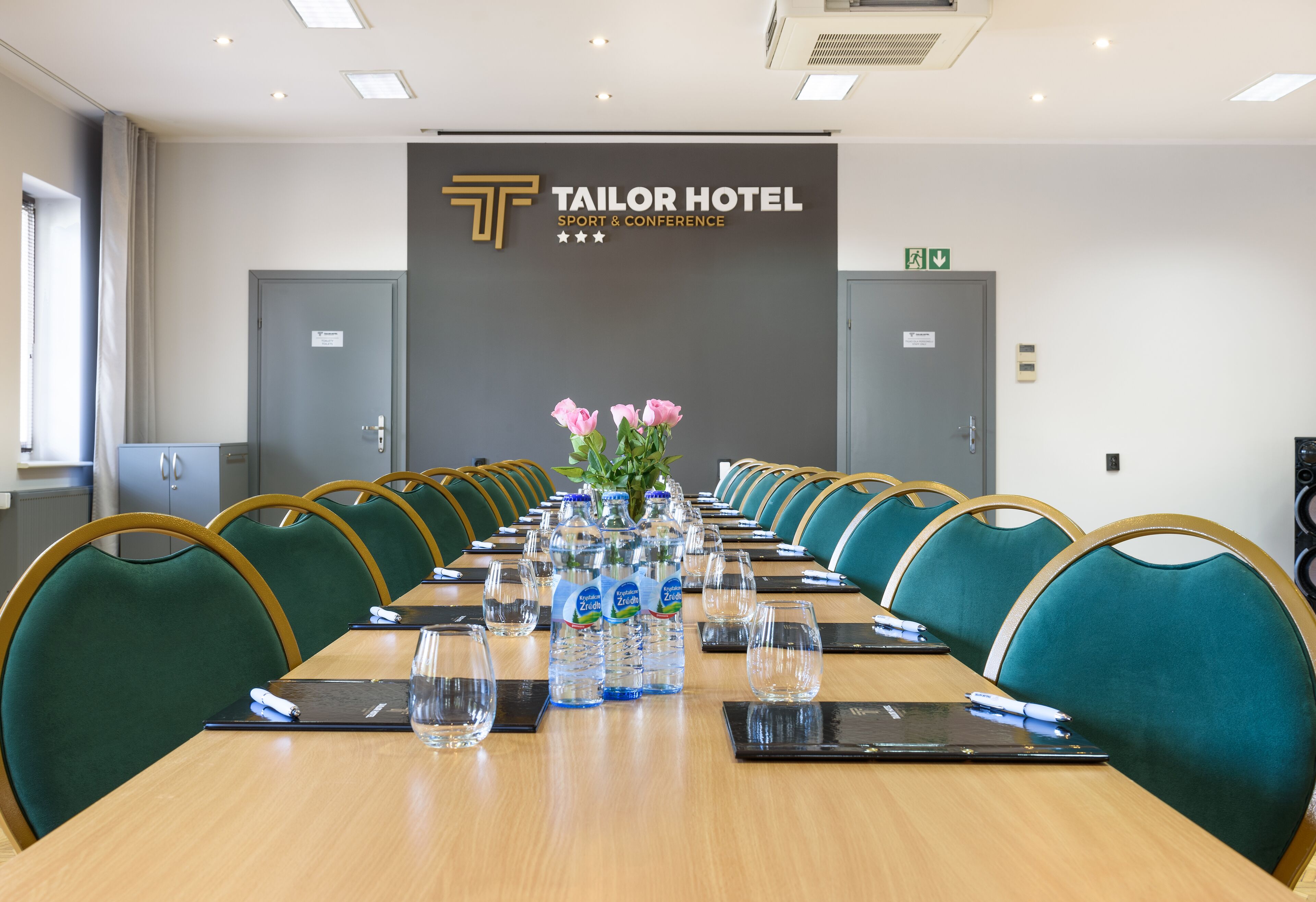 Una sala de conferencias en el Tailor Hotel con una larga mesa, sillas verdes, botellas de agua y un cartel que indica que es un espacio para deportes y conferencias.