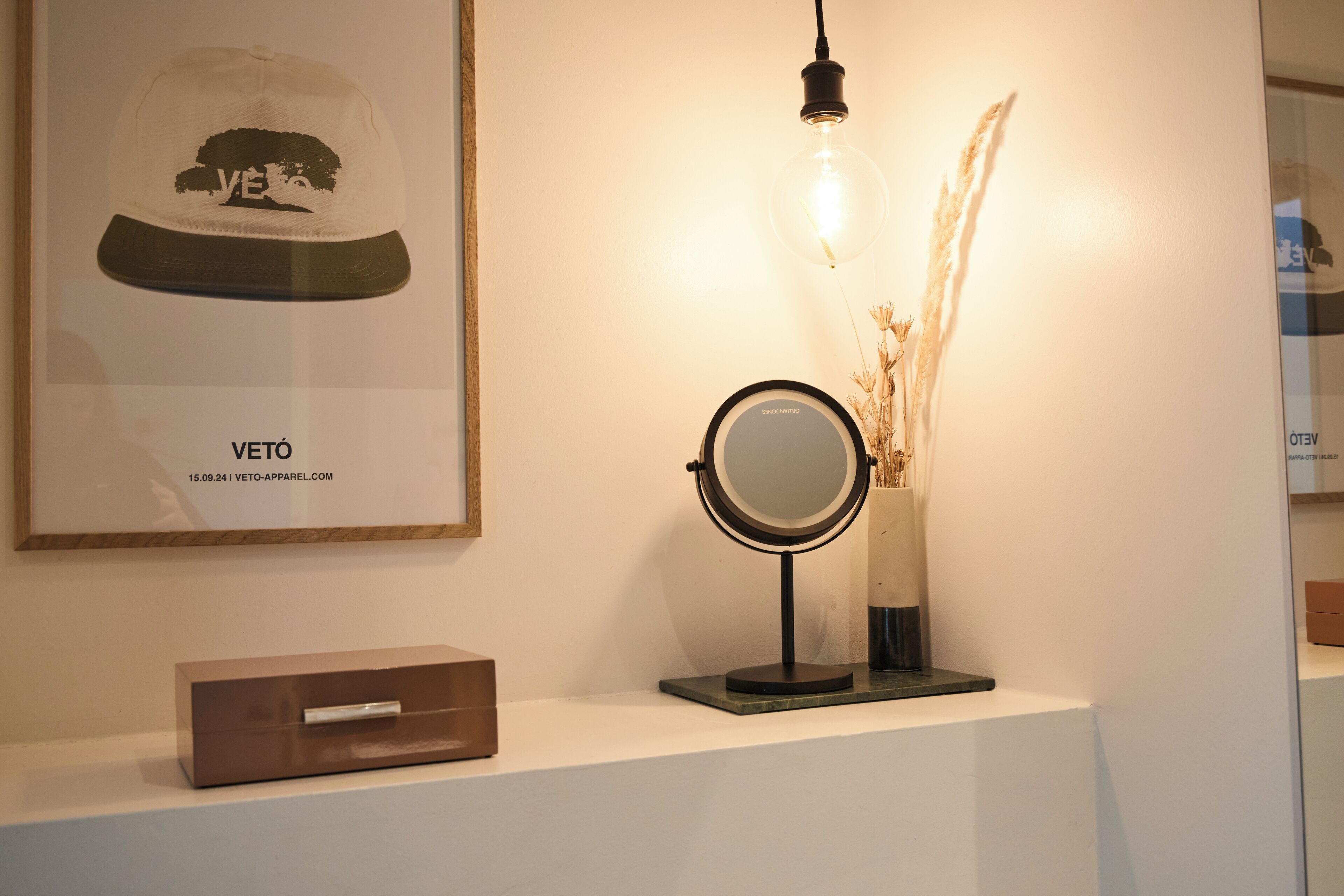 Une casquette Veto encadrée, un miroir rond et un vase contenant des plantes séchées, le tout posé sur une étagère blanche.