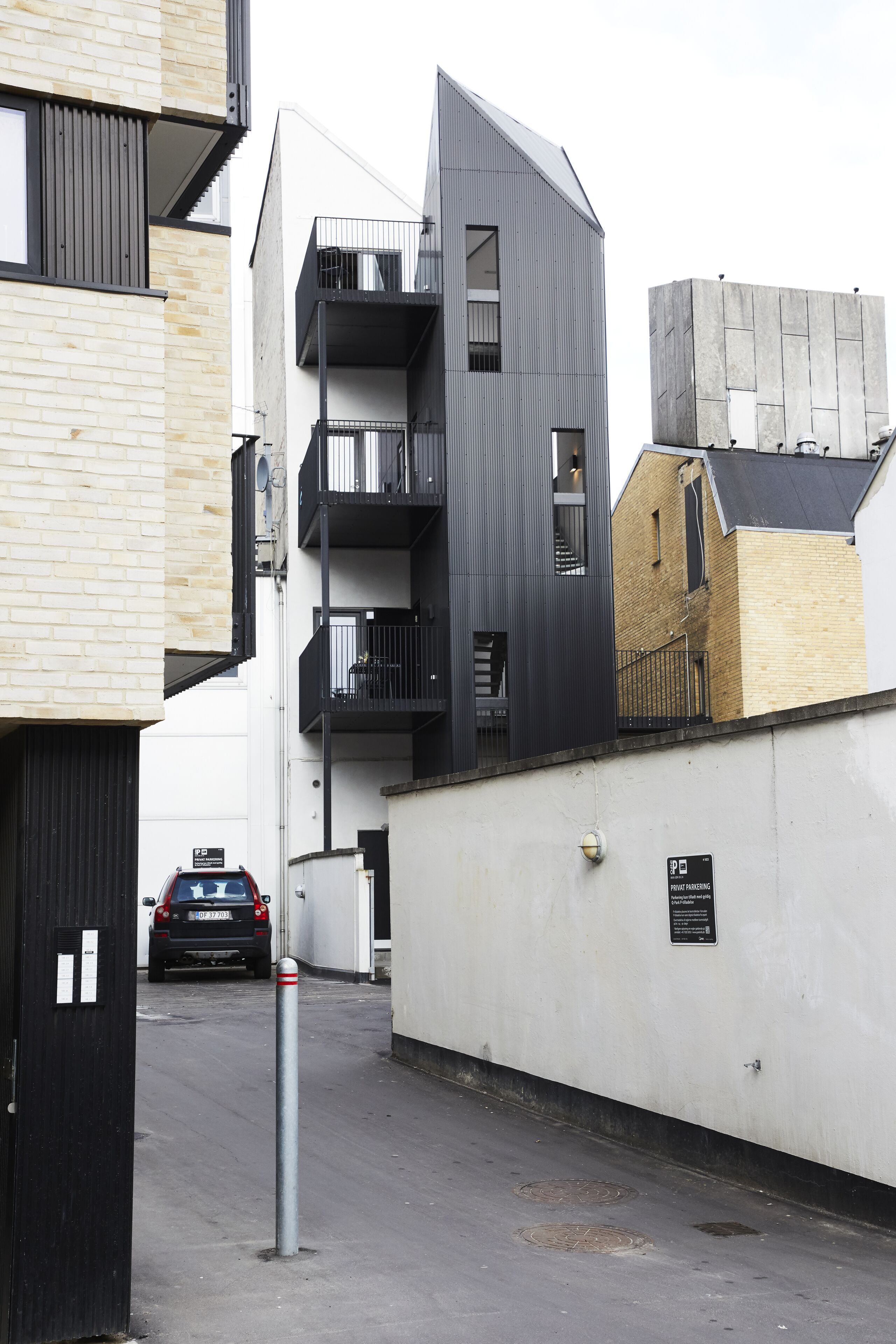 Une rue urbaine moderne, bordée d’un mélange de bâtiments résidentiels et commerciaux, avec notamment un panneau indiquant un parking et des bollards.