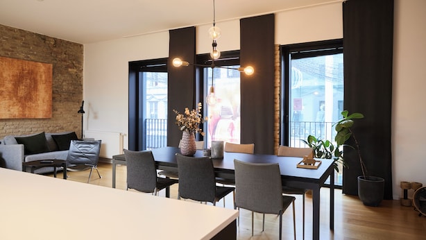 A modern dining area with a long table, multiple chairs, a vase with flowers, and a large window with curtains. - Søndergade - Strøget (Aarhus)