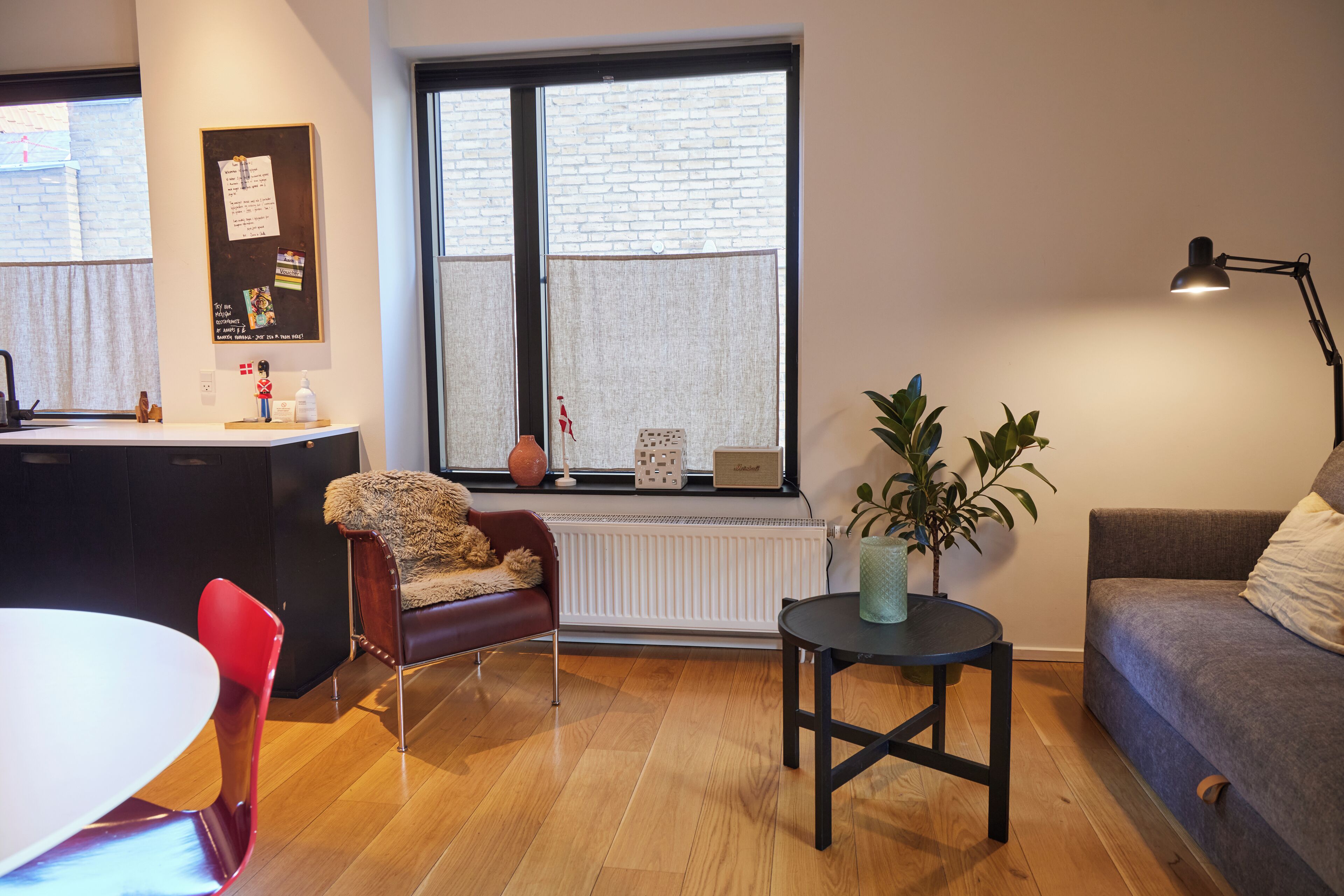 A modern living room with a dining area, a sofa, a small table with a plant, and a window with a view of a brick wall.