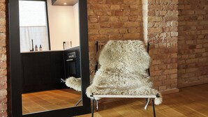A room with a brick wall, a wooden floor, a fluffy chair, and a mirror reflecting a kitchen area. - Amaliegade 10 B (Aarhus)