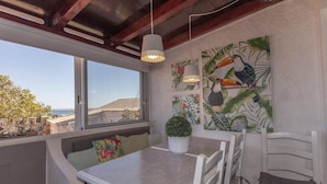 Zona pranzo con tavolo e sedie, vista mare attraverso una finestra e un murale a tema tropicale.
