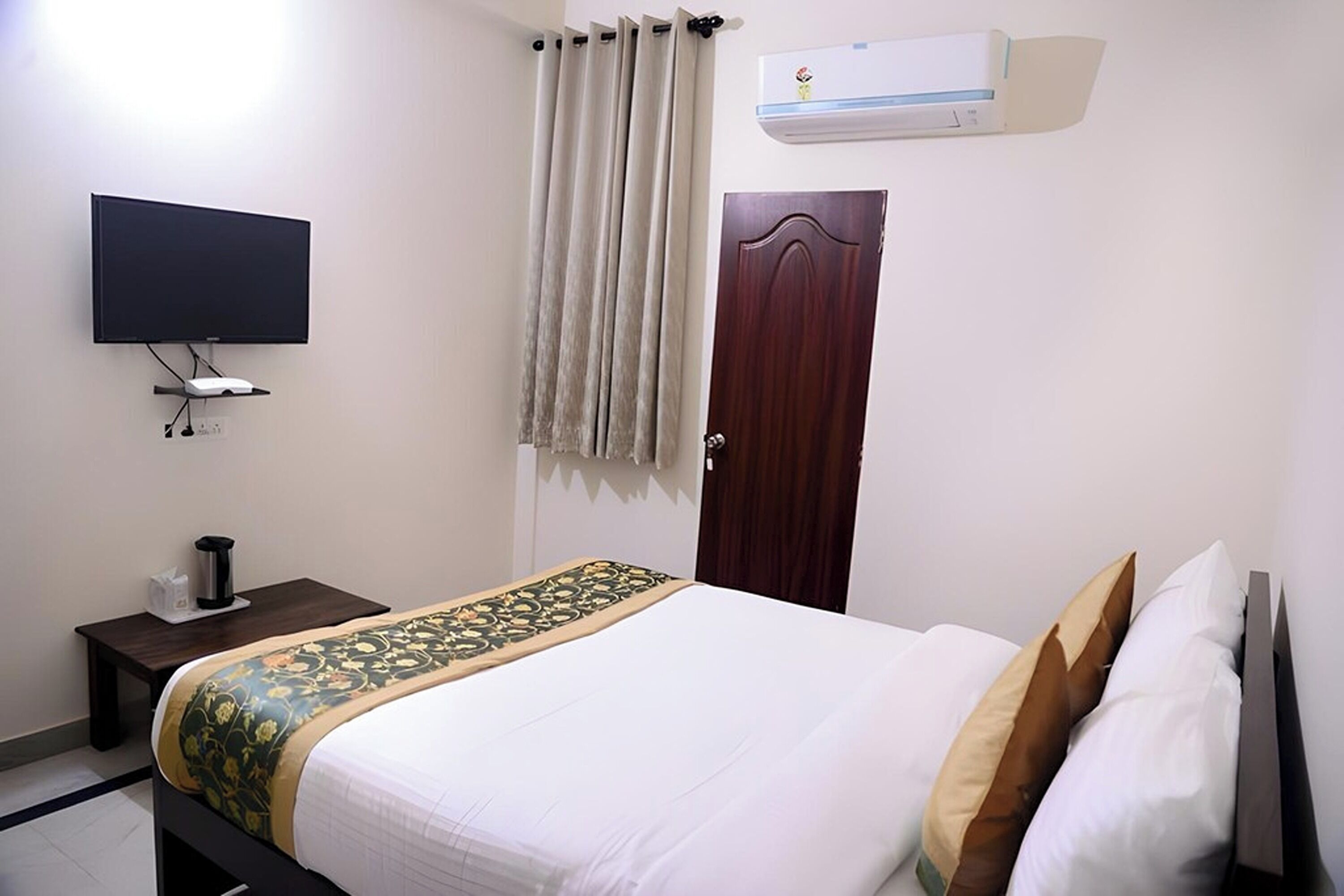 A hotel room with a bed, a television, a bedside table with a lamp, a curtain, and an air conditioning unit.