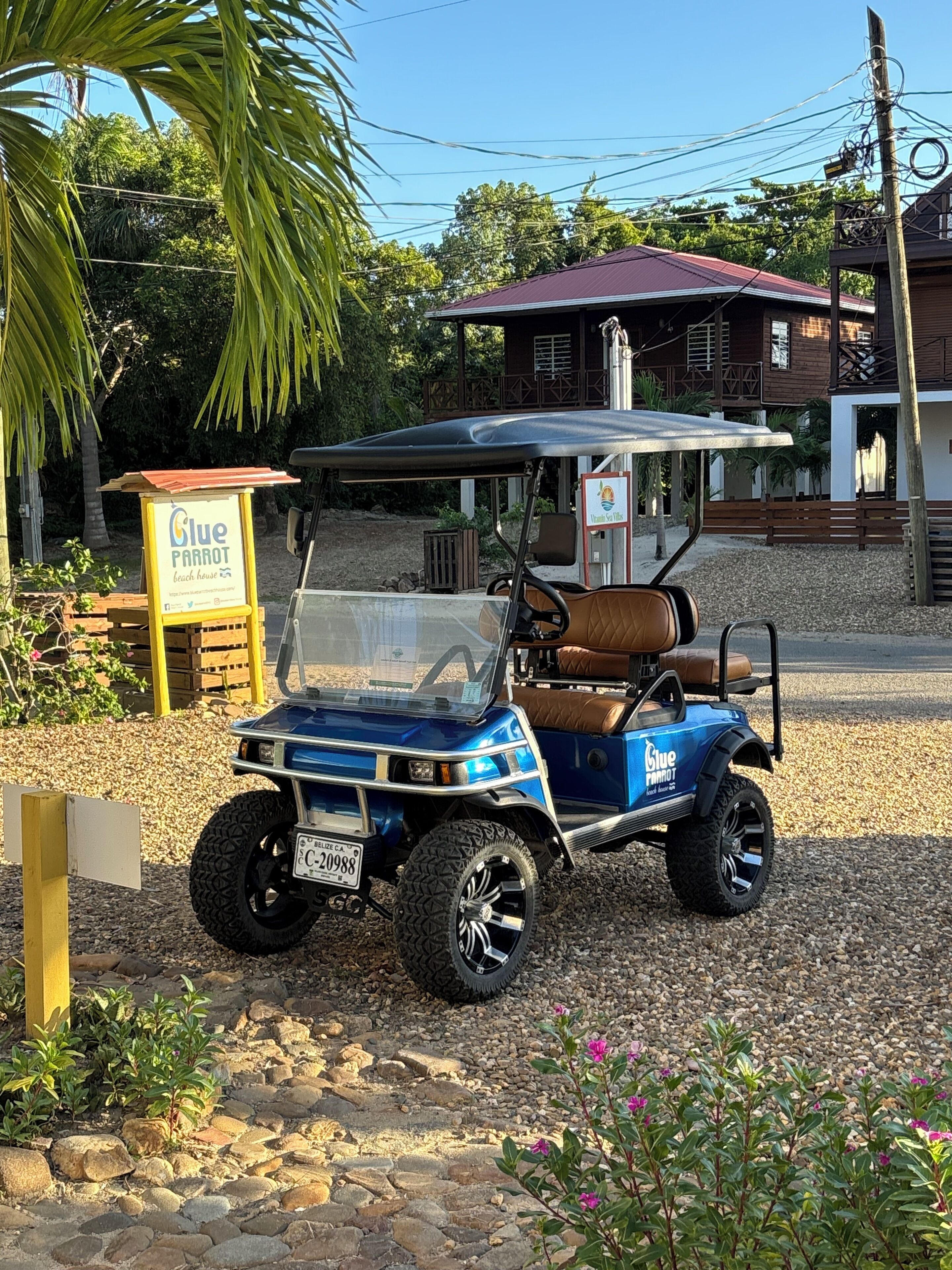 Une voiturette de golf bleue est garée sur un chemin de gravier, et on aperçoit en arrière-plan une enseigne indiquant « Glue Parrot Beach House ».