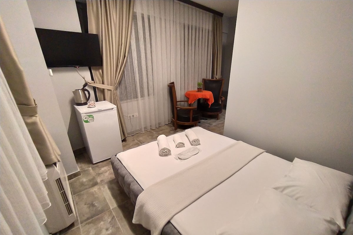 A hotel room with a bed, a desk with a kettle, a chair, and a TV on the wall.