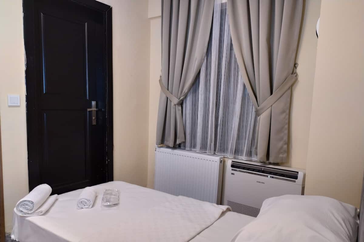 A small, neatly made bed with a white sheet and pillow, a rolled towel, and a glass container, likely for toiletries. A black door is visible to the left, and a radiator with a Samsung brand label is on the wall to the right.