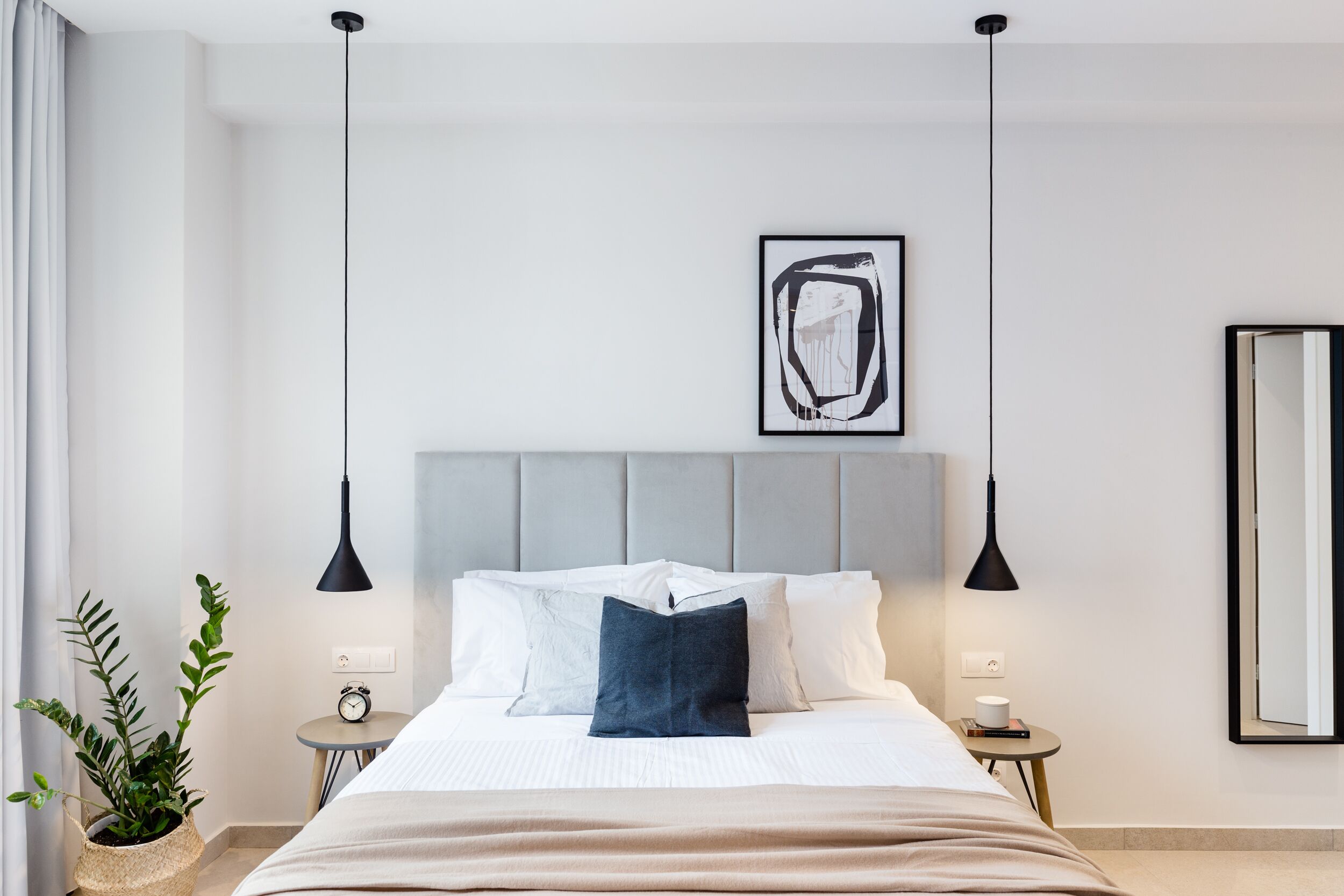 Une chambre d’hôtel moderne avec un grand lit, des tables de chevet, une plante en pot, une horloge murale et une œuvre d’art encadrée.