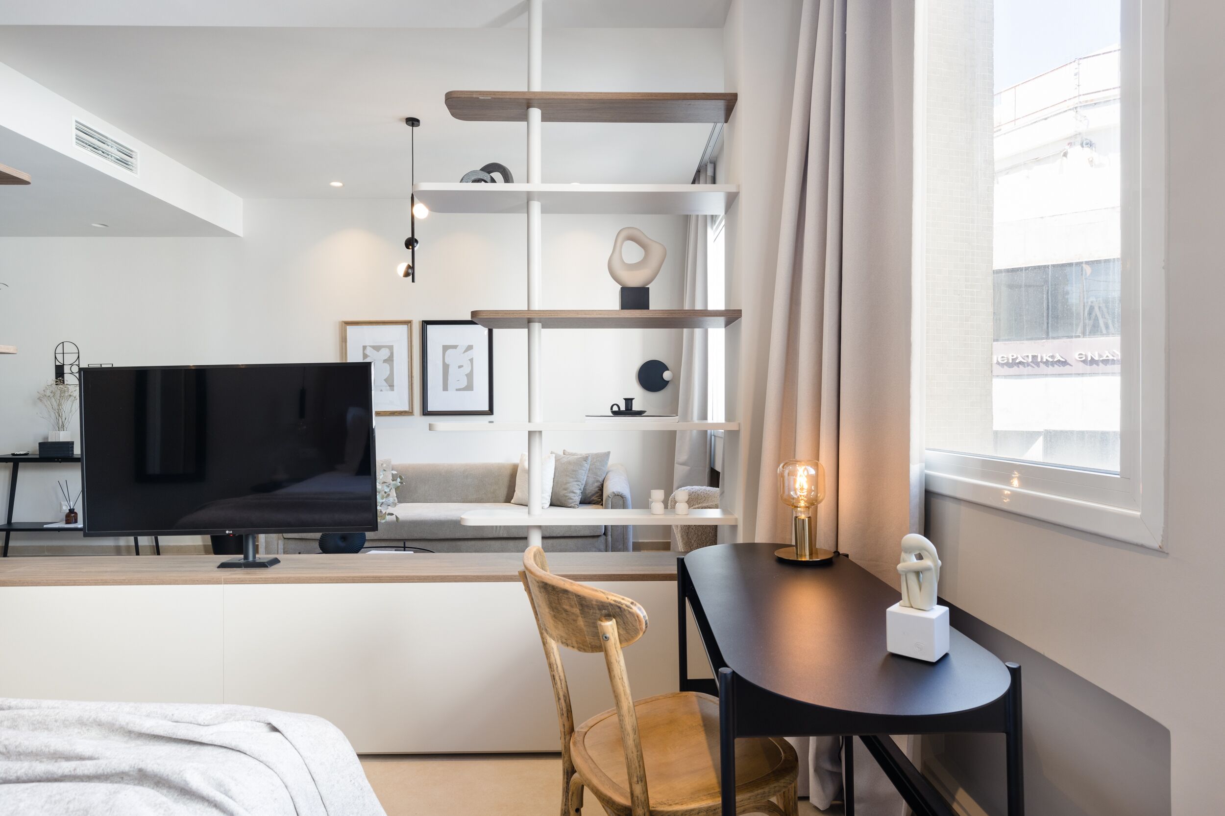 A modern hotel room with a flat-screen TV, a wooden chair, a small table with a sculpture, and a large window with a view of the city.