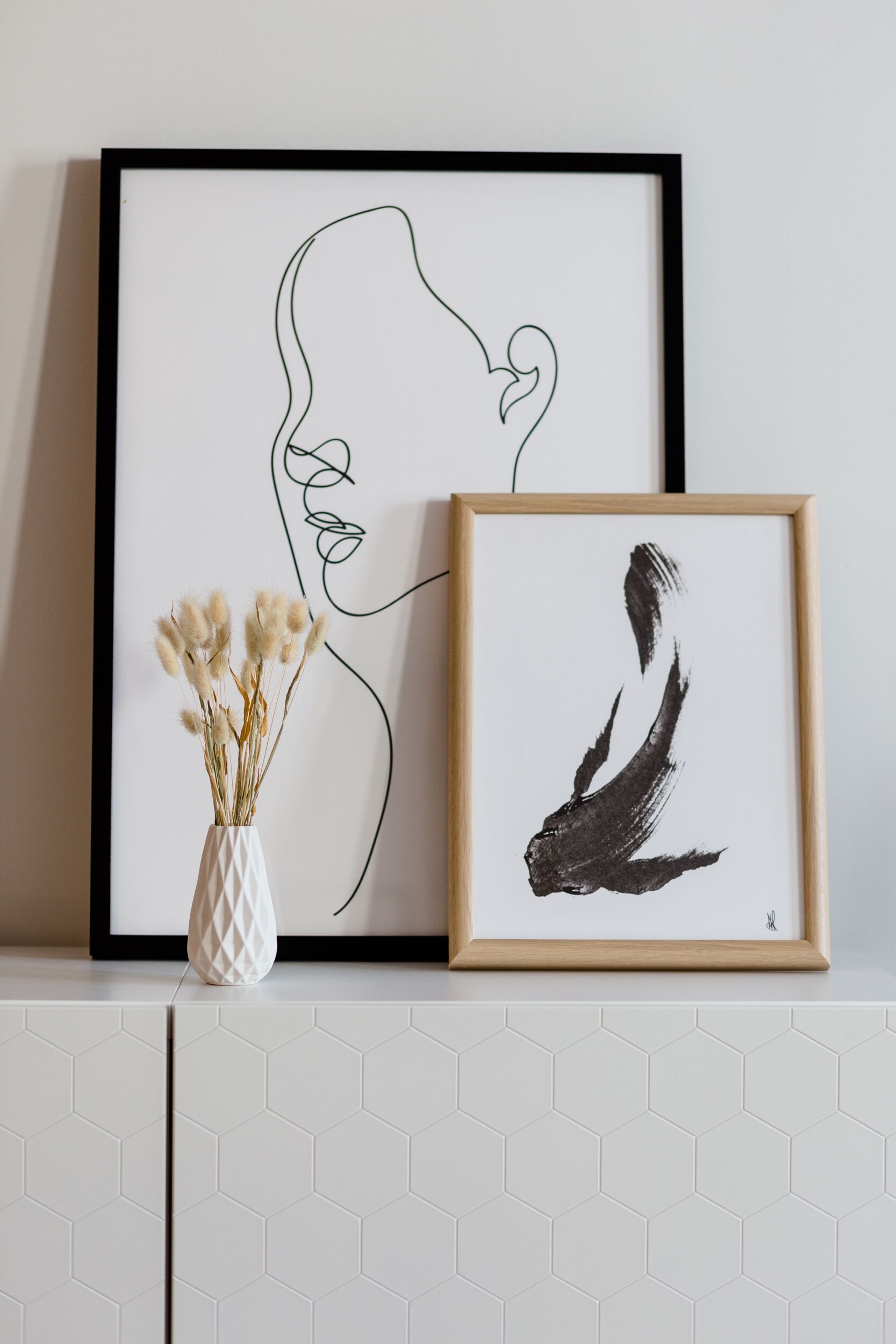 A framed line drawing of a woman's profile on a wall, a framed abstract painting, and a vase with dried flowers on a tiled surface.