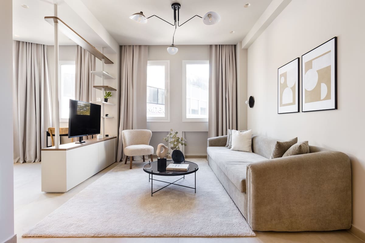 A modern living room with a flat-screen TV, a round coffee table, a beige sofa, and a white armchair. There are two windows with curtains, a ceiling light fixture, and two framed pictures on the wall.
