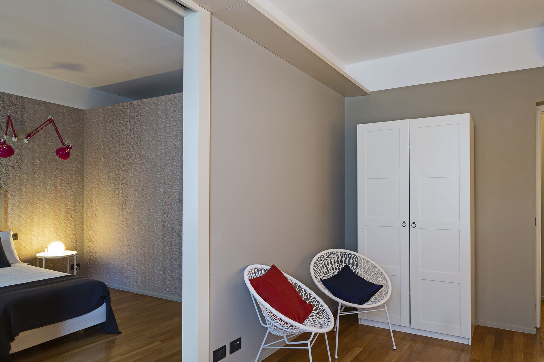 Una stanza con un letto, due poltrone in vimini bianco con cuscini rossi e blu, un armadio bianco e una parete decorata.