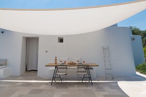 An outdoor dining area with a table, two chairs, a ladder, and a white wall.