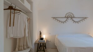 Un dormitorio con una cama, estante de madera con ropa, escritorio con lámpara y un adorno en la pared.