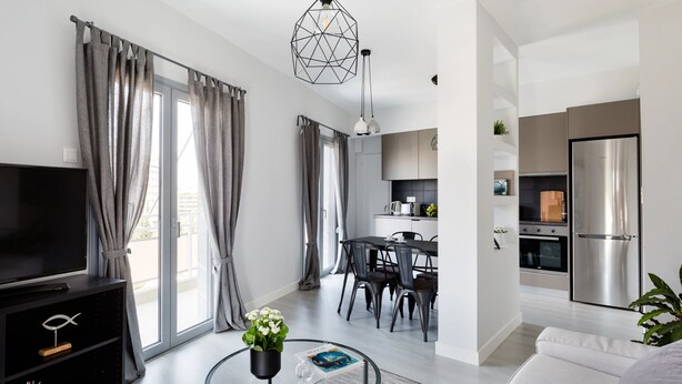 A modern living room with a flat-screen TV, a dining area with a table and chairs, a kitchen with a refrigerator and stove, and large windows with curtains. - UPSTREET Minimal 2BD Apt-Lycabettus view (Kaisariani)