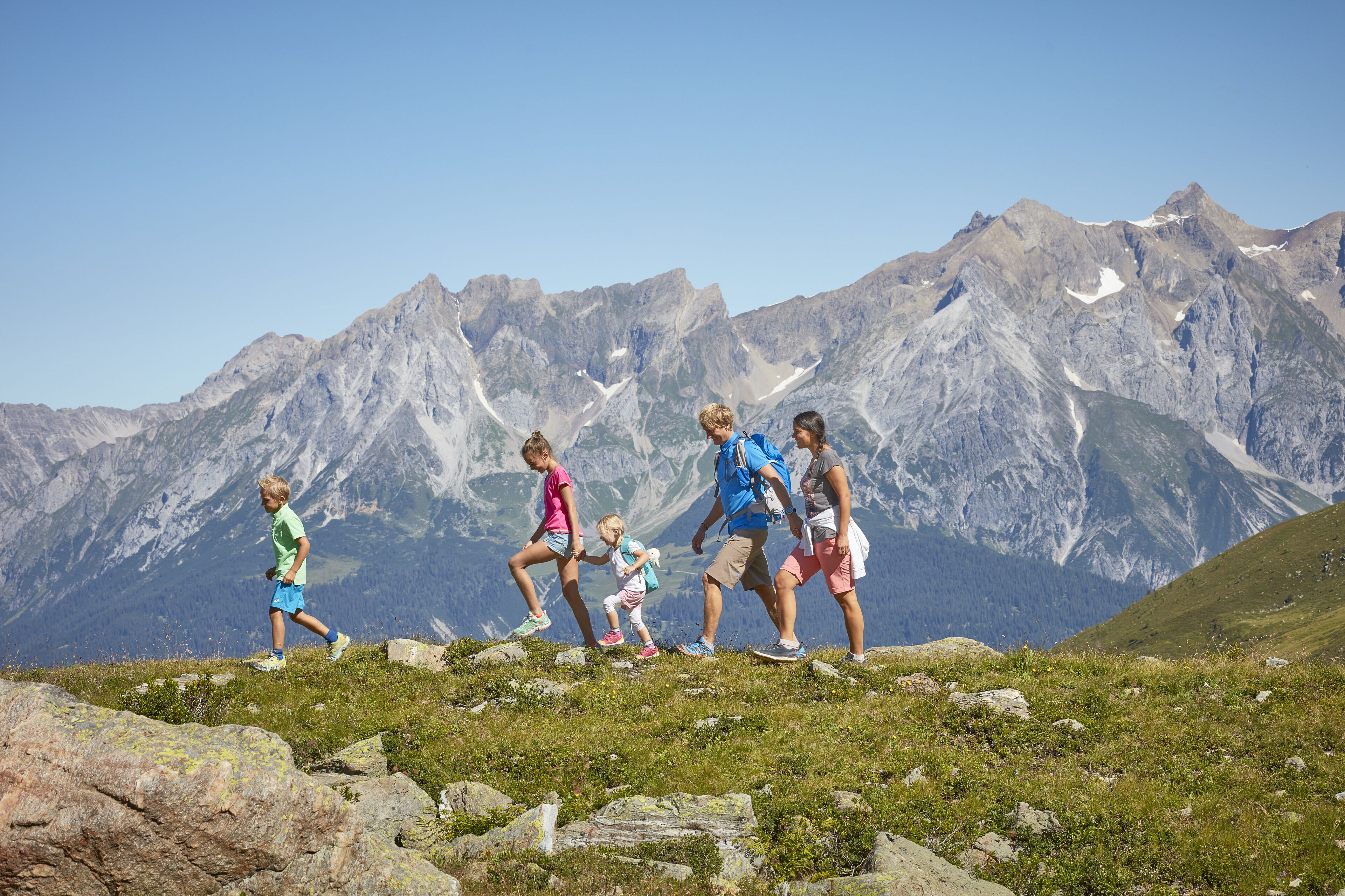 Eine Gruppe Wanderer auf einem grasbewachsen Bergpfad mit felsigem Untergrund und einer Bergkulisse.