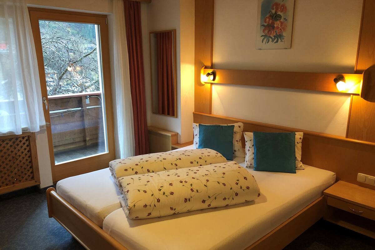 A hotel room with a bed, a nightstand, a bedside table, a window with curtains, and a painting on the wall.