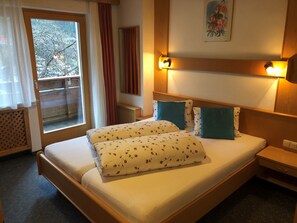 A hotel room with a bed, a nightstand, a bedside table, a window with curtains, and a painting on the wall. - Hotel Garni Bergwelt (See)