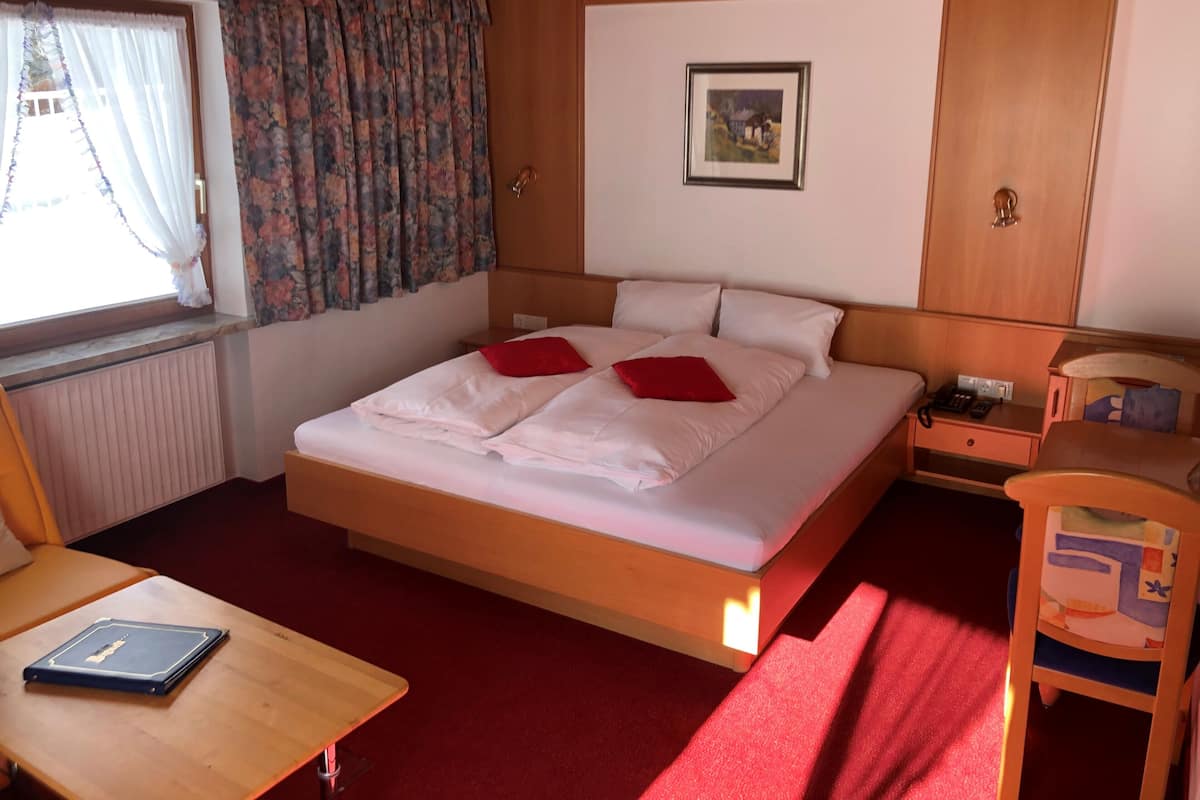 A neatly arranged hotel room with a large bed, a wooden headboard, a small bedside table with a lamp, a chair, a window with curtains, and a framed picture on the wall.