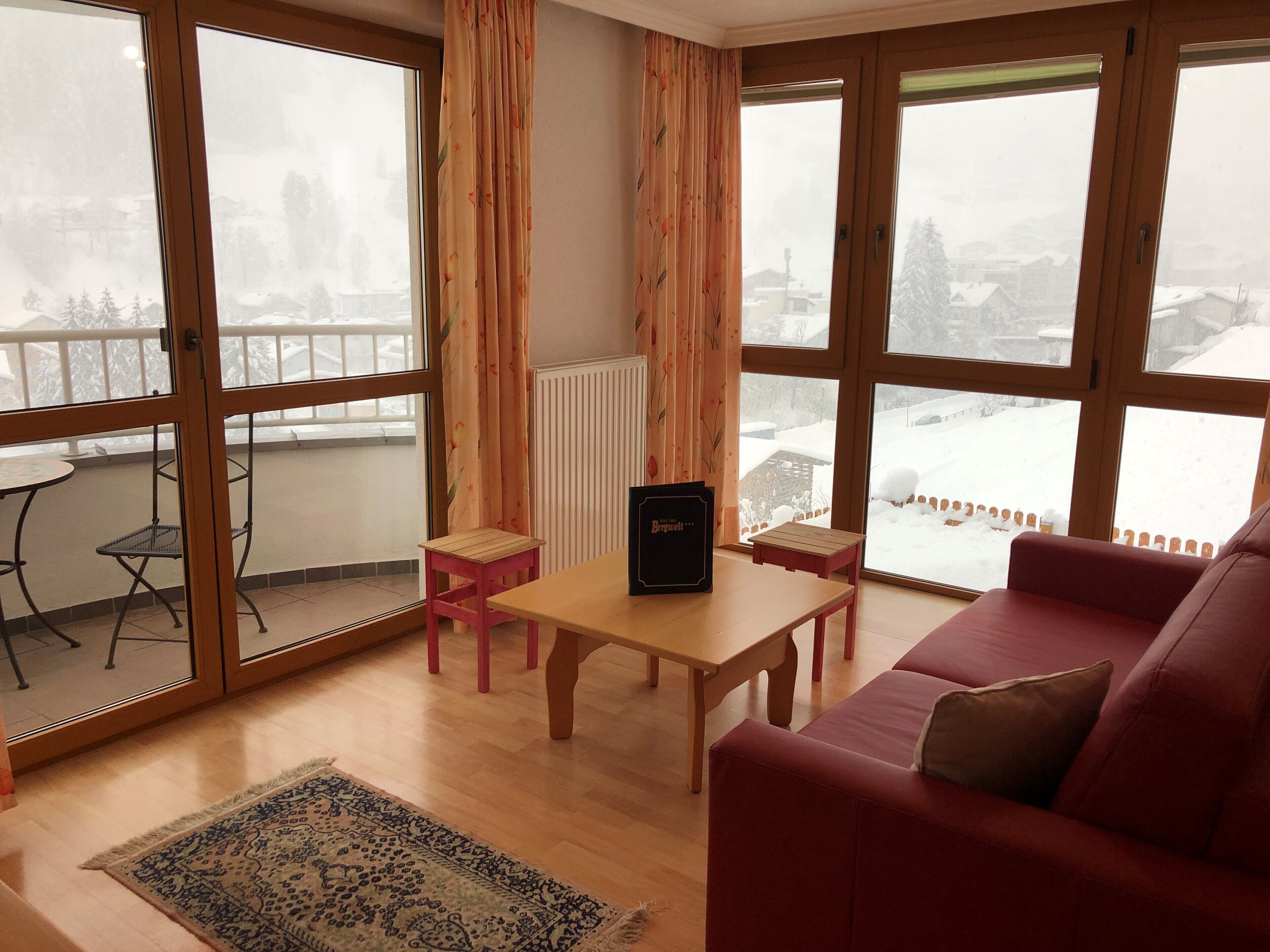 A cozy living room with a red sofa, a wooden coffee table, a small table, and a chair. There is a large window with a view of a snowy landscape.