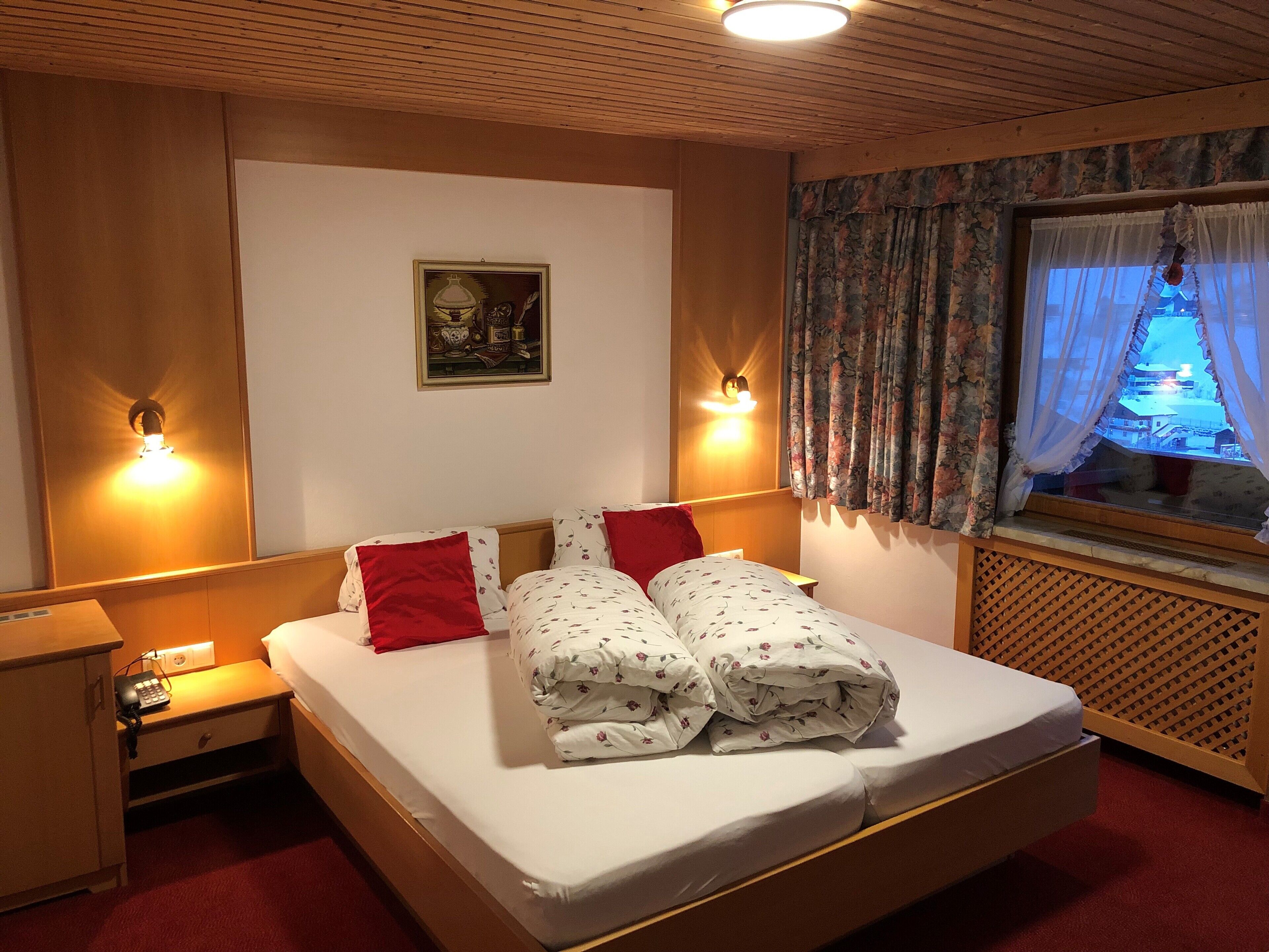 Una habitación de hotel con cama, dos mesitas de noche, una lámpara de pared, ventana con cortinas e imagen enmarcada en la pared.