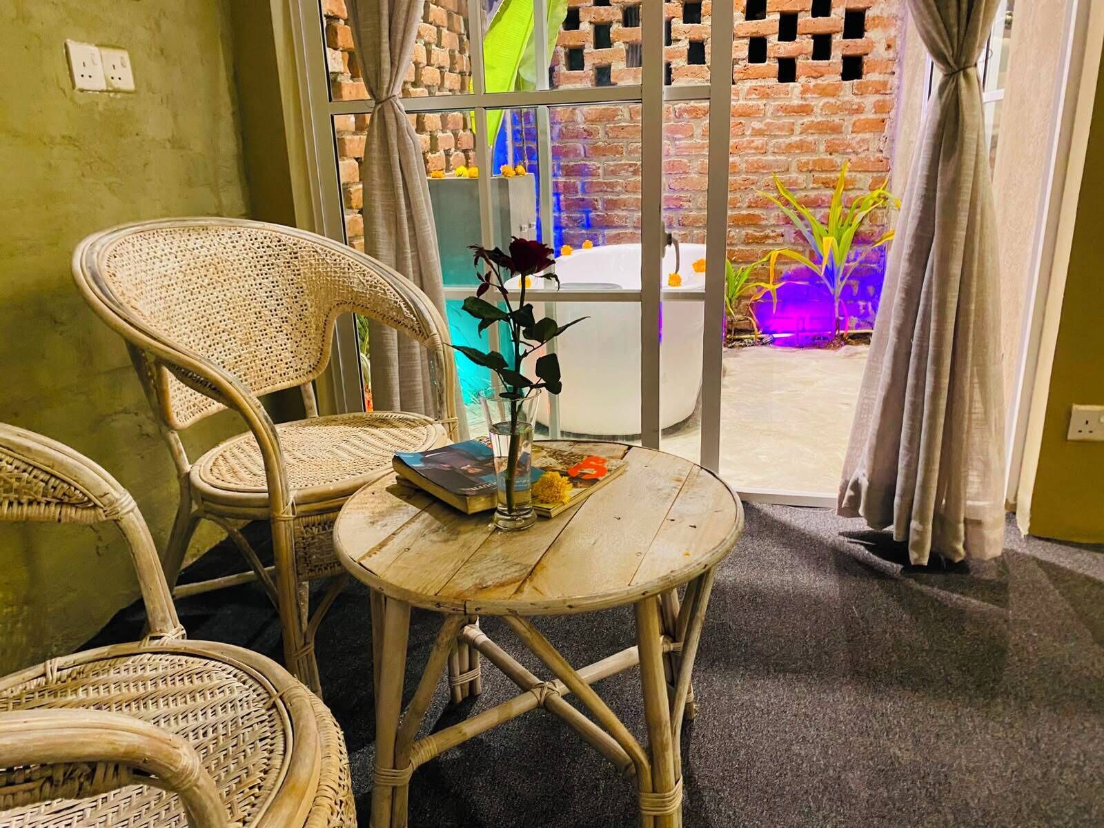 Un ensemble de chaise et de table en osier est installé près d’une porte vitrée, offrant une vue sur un mur de briques et une piscine.