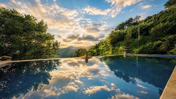 Ein klarer Blick auf ein Schwimmbecken mit einer Person, die am Rand entspannt, umgeben von üppiger Vegetation und Bergen im Hintergrund.