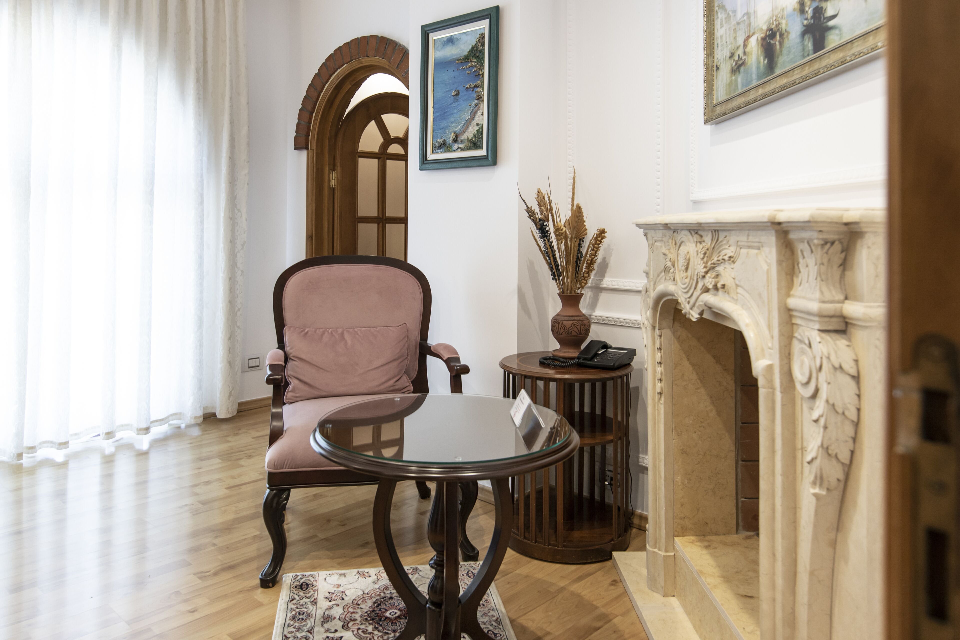 Una habitación con piso de madera, un sillón rosa, una mesa de vidrio, una pequeña mesa redonda con un florero y un teléfono, una chimenea y un cuadro en la pared.