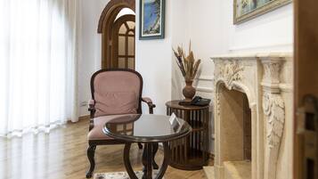 Una habitación con piso de madera, un sillón rosa, una mesa de vidrio, una pequeña mesa redonda con un florero y un teléfono, una chimenea y un cuadro en la pared.
